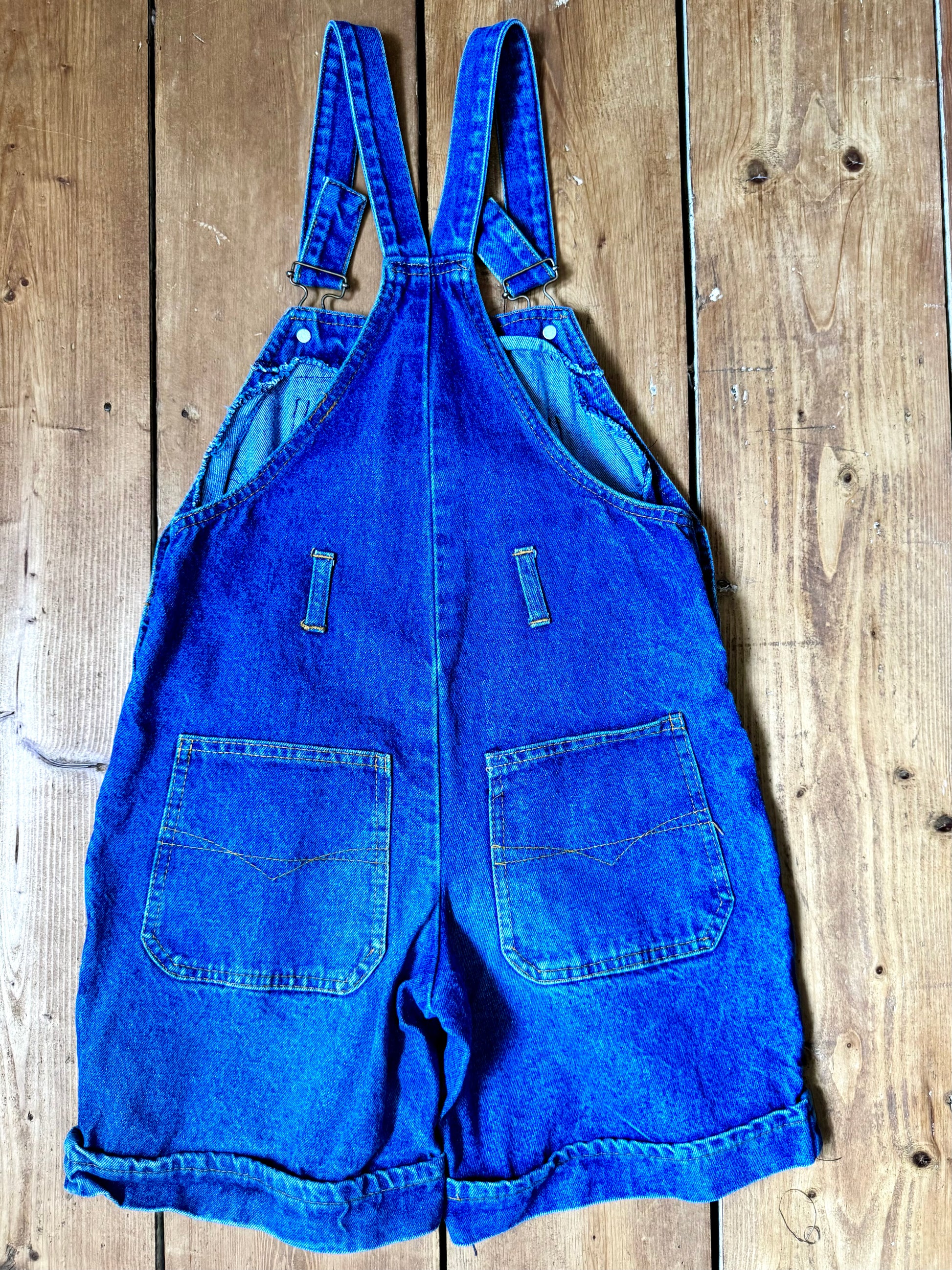 Vintage blue denim dungaree shorts for kids with embroidered chest detail and adjustable straps. Authentic late 80s/early 90s, handpicked for Bee Tiny kids vintage.