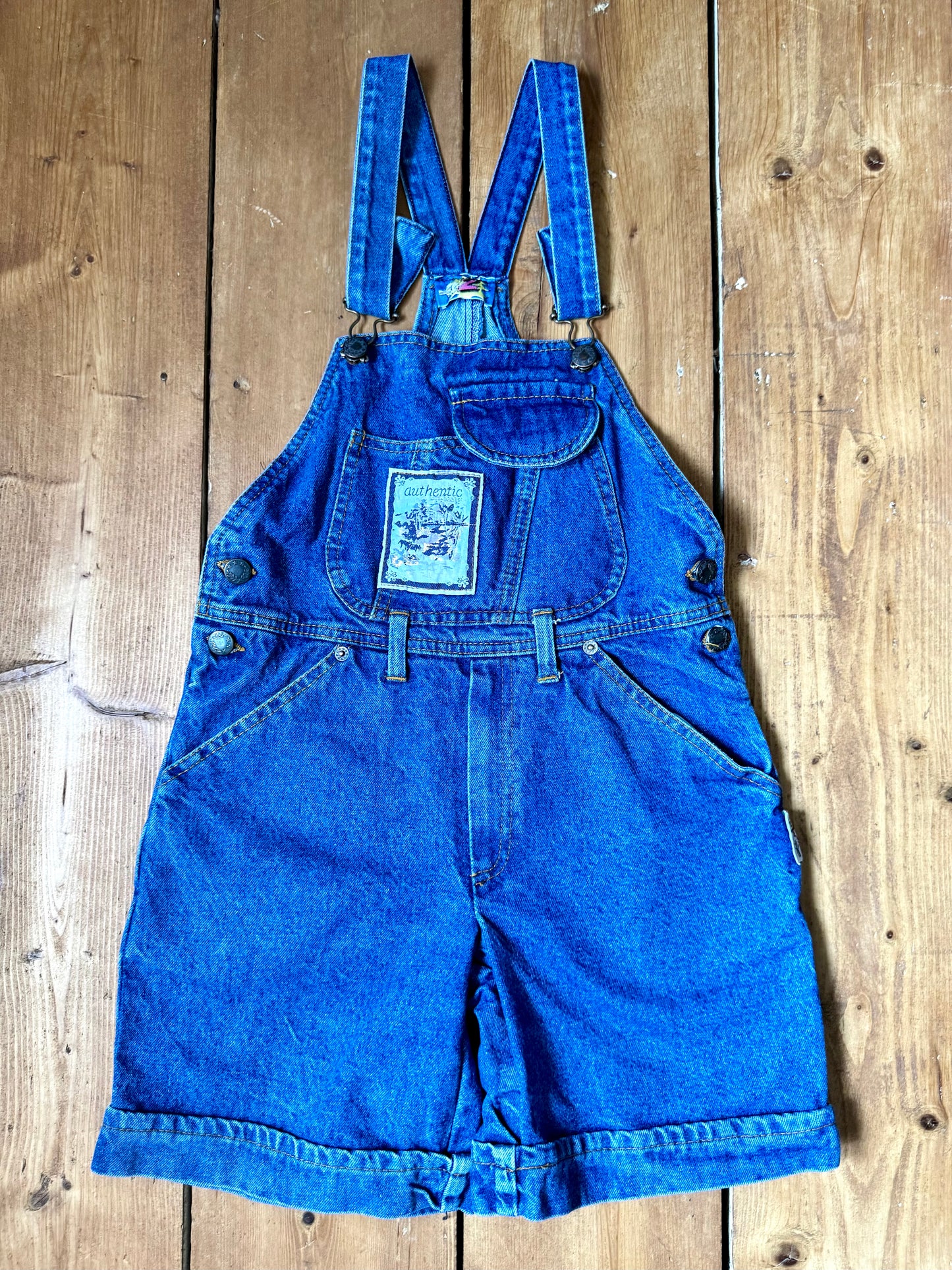 Vintage blue denim dungaree shorts for kids with embroidered chest detail and adjustable straps. Authentic late 80s/early 90s, handpicked for Bee Tiny kids vintage.