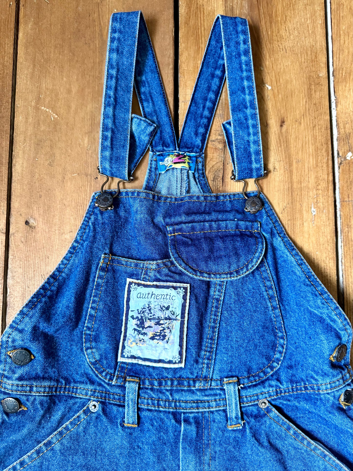 Vintage blue denim dungaree shorts for kids with embroidered chest detail and adjustable straps. Authentic late 80s/early 90s, handpicked for Bee Tiny kids vintage.